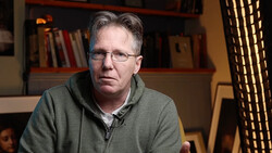 Man wearing glasses and gray hoodie sitting indoors with bookshelves in background.