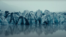 Jagged glacier face with dark crevasses reflected in still water below.