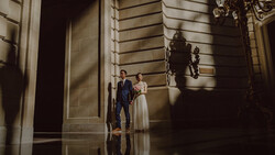 Bride and groom standing in a classical hallway with dramatic side lighting casting shadows on stone walls.