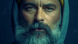 Bearded man with intense gaze wearing a teal hood against dark background.
