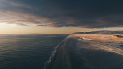 Aerial view of a sandy coastal spit extending into dark ocean waters at dusk.