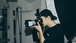 Photographer in black cap and shirt holding a professional video camera with yellow shoulder strap in a studio setting.
