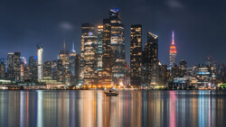 Illuminated Manhattan skyline at dusk reflected in calm river water.