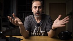 Man in a dark v-neck shirt gesturing expressively while speaking to camera indoors.