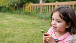 Young girl in pink shirt looking down at smartphone in backyard setting.