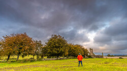 Solitary figure in bright orange jacket standing in open green field beneath dramatic overcast sky.