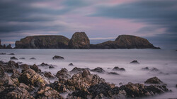 Rocky shoreline with smooth water and three distant islands under a twilight sky with pink and blue clouds.