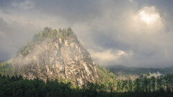 Dramatic rocky peak surrounded by dense forest and swirling mist under cloudy sky.