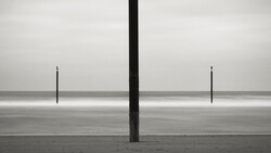 Three vertical posts on a vast, flat beach with calm water and overcast sky in stark monochromatic composition.