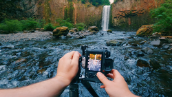 Photographer's hands holding a camera displaying a waterfall scene in the LCD screen, with the actual waterfall and rocky stream visible in the background.