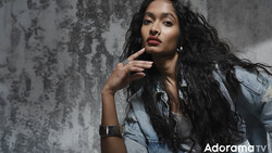 Woman with long dark hair posing against a textured gray wall, hand raised to face.