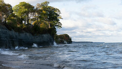 Rocky cliff shoreline with trees and breaking waves under cloudy sky.