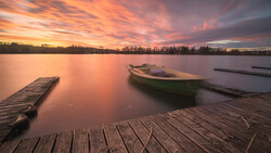 Wooden dock and moored boat on calm lake at sunset with dramatic orange and purple sky.