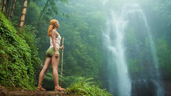 Woman standing on a forest trail gazing toward a misty waterfall in the distance.