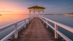 Wooden pier with white railings extending toward a gazebo at sunrise over calm water.
