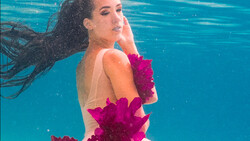 Woman underwater in turquoise pool wearing magenta ruffled sleeves, long dark hair flowing, looking toward camera.