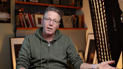 Man in gray zip-up hoodie and glasses speaking during an interview or presentation in an office setting.