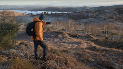 Hiker in orange jacket with backpack overlooking a valley with lakes and bare trees during golden hour.