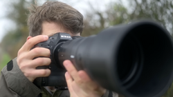 Photographer holding a Nikon DSLR with telephoto lens aimed at camera. Blurred background shows trees and foliage.