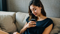 Woman in black shirt looking at smartphone while sitting on beige couch.