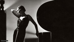 Woman in dark dress poses against geometric black shapes in high-contrast studio lighting.