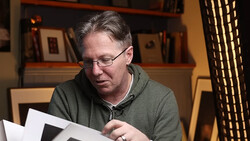 Man wearing glasses and olive hoodie reviewing contact sheets at a desk.