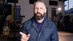 Bearded man in denim shirt gesturing while speaking in a professional studio setting.