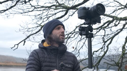 Videographer in winter gear operating a professional mirrorless camera on a tripod overlooking a winter landscape.