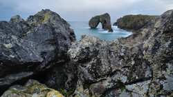 Rugged coastal rock formations with turquoise water and a natural stone arch in the distance.