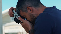 Photographer holding a mirrorless camera to his face in bright daylight conditions.