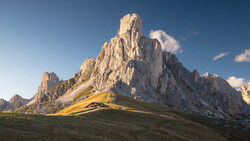 Dramatic limestone mountain peak with golden meadow in foreground under clear blue sky.
