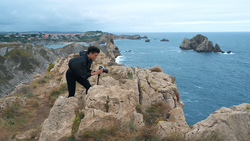 Photographer crouching on rocky coastal outcrop while framing a shot of distant sea stacks and turquoise waters.