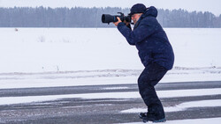 Photographer in dark winter coat crouched on frozen lake while shooting with telephoto lens, forest treeline visible across snowy landscape.