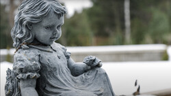 Stone statue of a young child in profile, holding a small flower, with weathered patina.