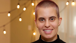 Portrait of a young person with short reddish-brown hair wearing a black turtleneck, smiling at the camera against a warm-lit background with bokeh lights.