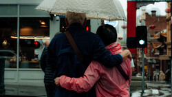 Three people from behind holding an umbrella in rainy urban setting with storefronts visible.