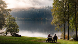Two people sitting at a picnic table on a grassy shore, facing a calm lake surrounded by evergreen forest under misty mountains.