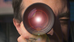 Close-up of a photographer's face looking through a camera lens with a bright red glow and internal reflections visible.