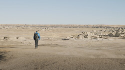 Solitary hiker with blue backpack walking across vast, barren desert terrain scattered with rocks.