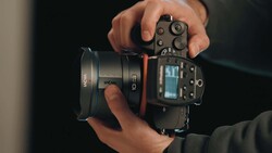 Photographer's hands holding a mirrorless camera with attached lens against a dark background.
