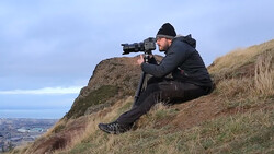 Photographer in dark jacket crouched on hillside aiming telephoto lens toward distant landscape.