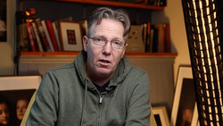 Man wearing glasses and olive hoodie speaking in front of bookshelf.