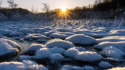 Icy boulders scattered across a partially frozen river at sunrise, backlit by golden sunlight and bare trees.