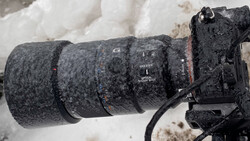 Telephoto lens covered in frost and ice mounted on camera body in snowy conditions.