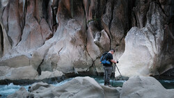 Photographer with tripod standing on ice formations beneath towering white and gray cliff walls.