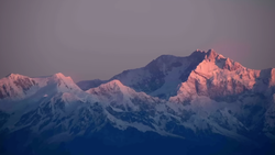 Snow-covered mountain range illuminated by alpenglow at sunrise or sunset, with pink and warm light on peaks against a cool blue sky.