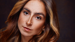 Portrait of a woman with long reddish-brown hair and freckles against a dark background.