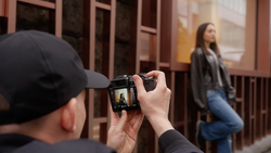 Photographer in black cap holding compact camera while framing shot of woman in doorway.