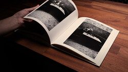 Hand holding open a photography book displaying black and white landscape images on a wooden table.