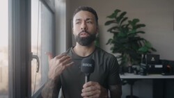 Male content creator speaking into a microphone while gesturing toward a window in a studio setting.